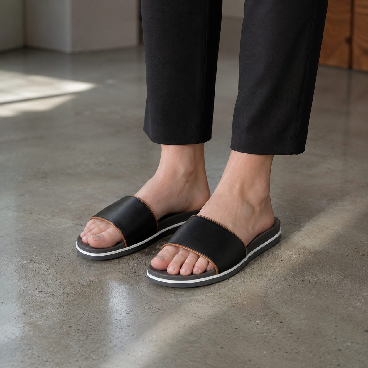 Woman wearing matte black single-band vegan leather slide sandals with cropped black trousers on a polished concrete loft floor