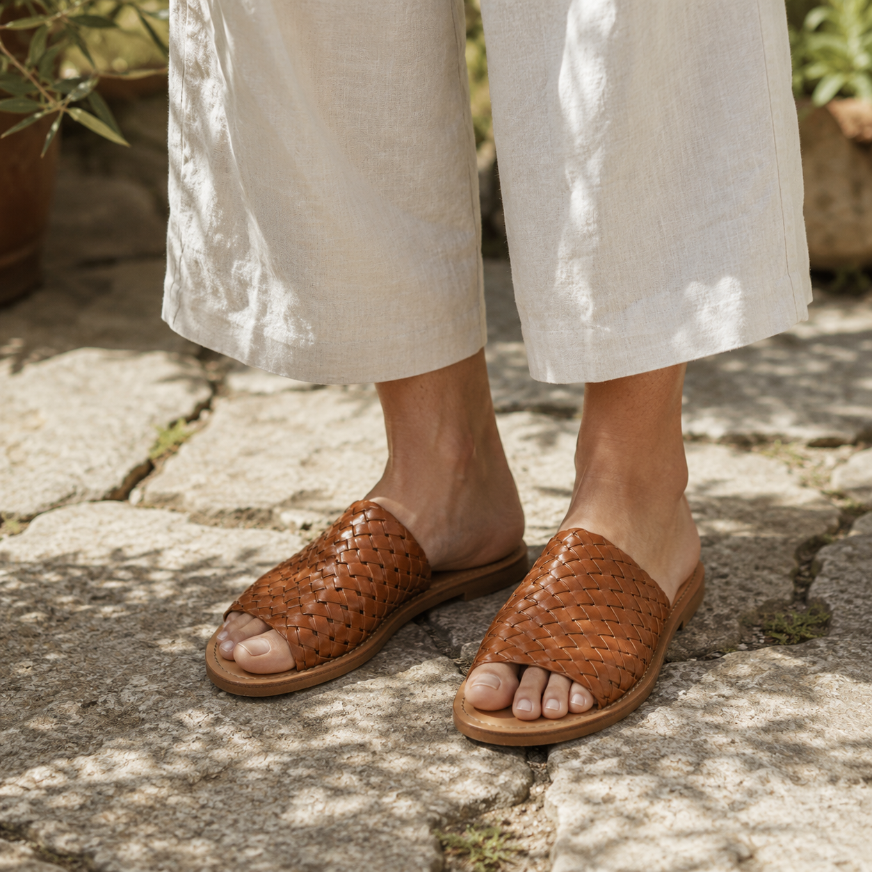Woman wearing cognac hand-woven vegan leather slide sandals with cream linen wide-leg trousers on a sun-warmed Mediterranean stone garden path